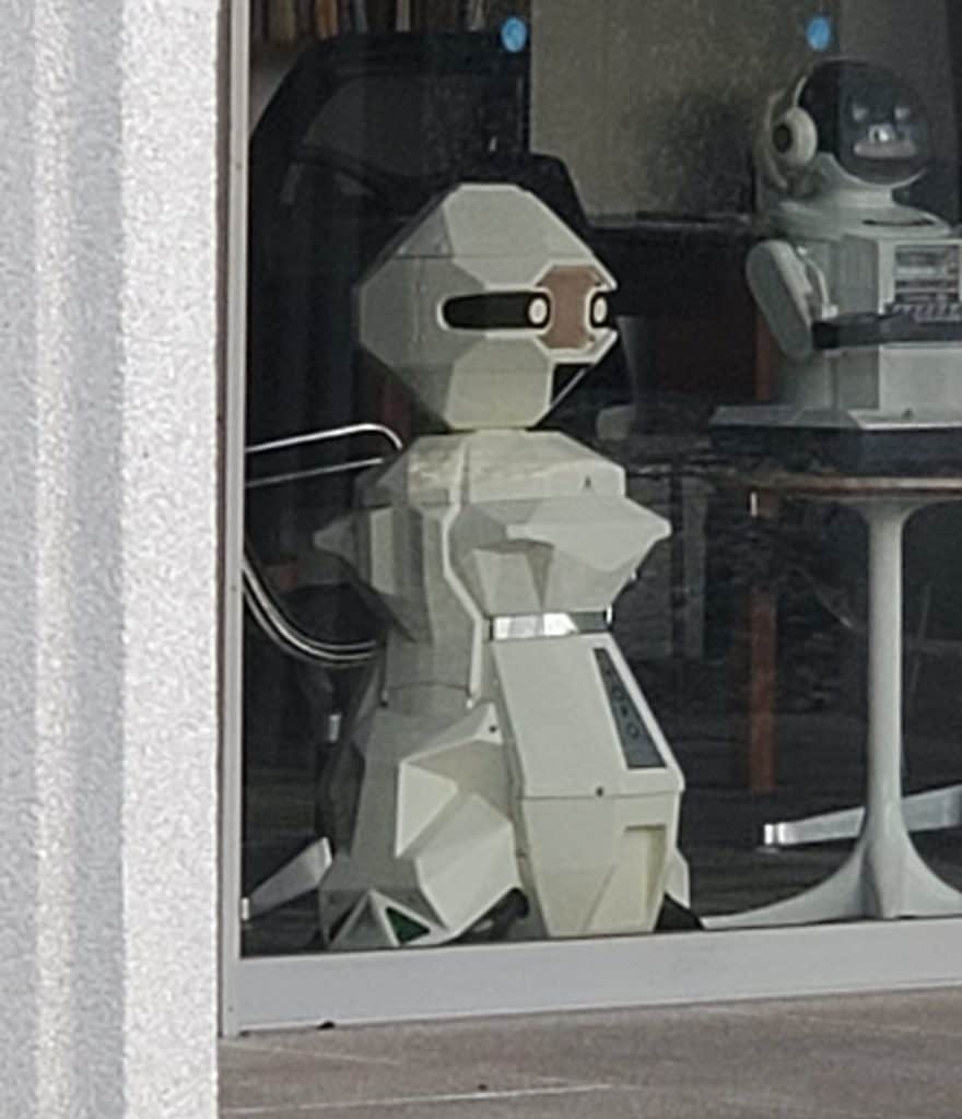 Side view of a white TOPO robot with a geometric design and black visor, placed next to another retro robot.